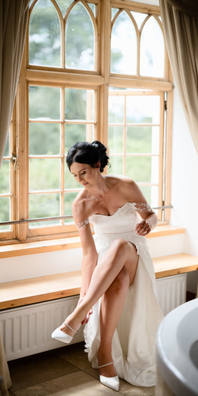 Bride putting on her shoes while sitting by a window