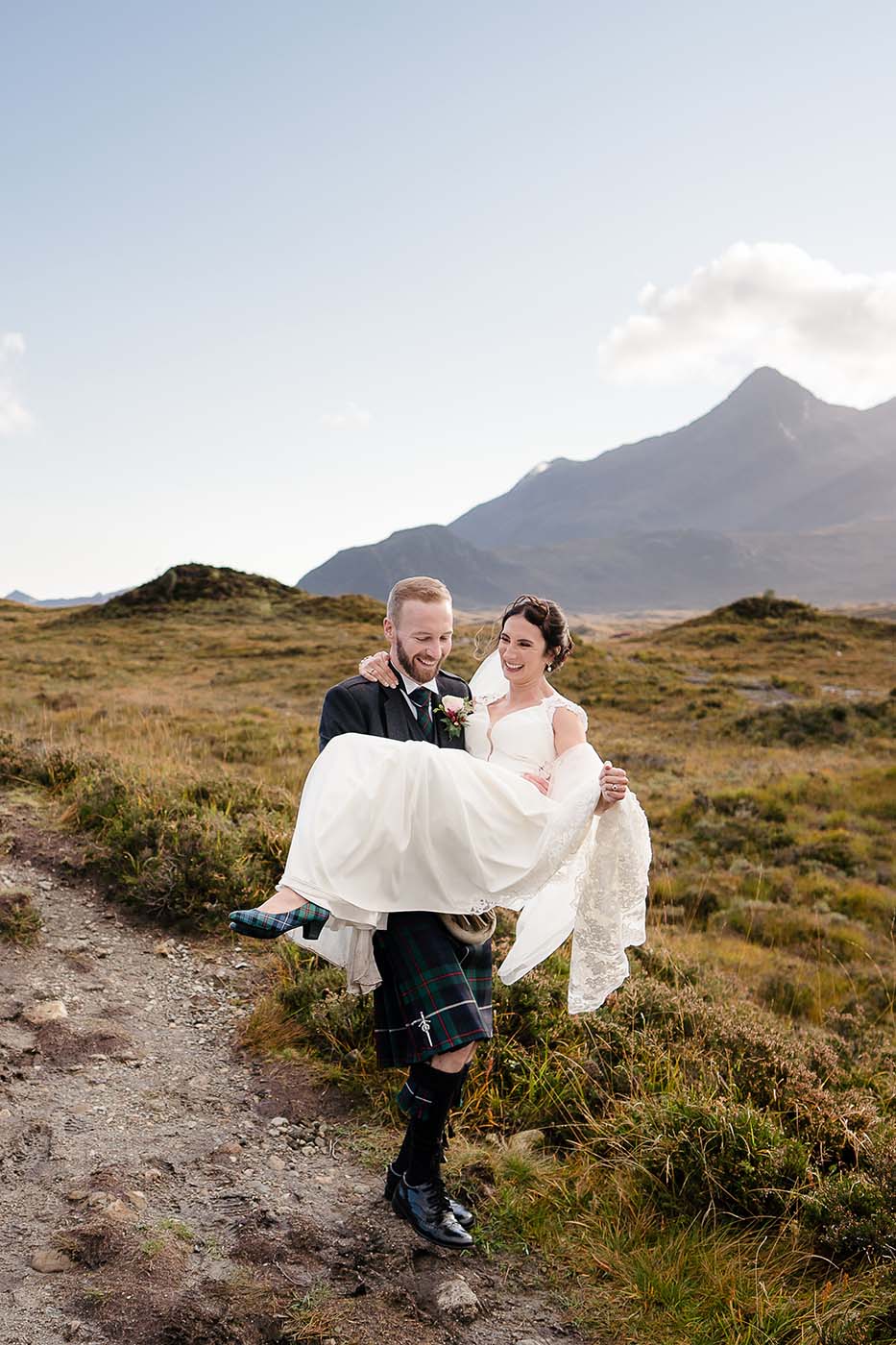 Groom carrying his bride on hillside path with cuillin mountains isle of skye in background