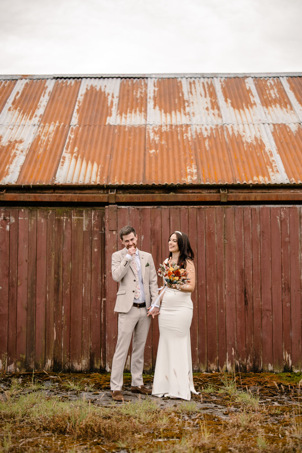 Bride and groom laughing together next to an old shed on their wedding day