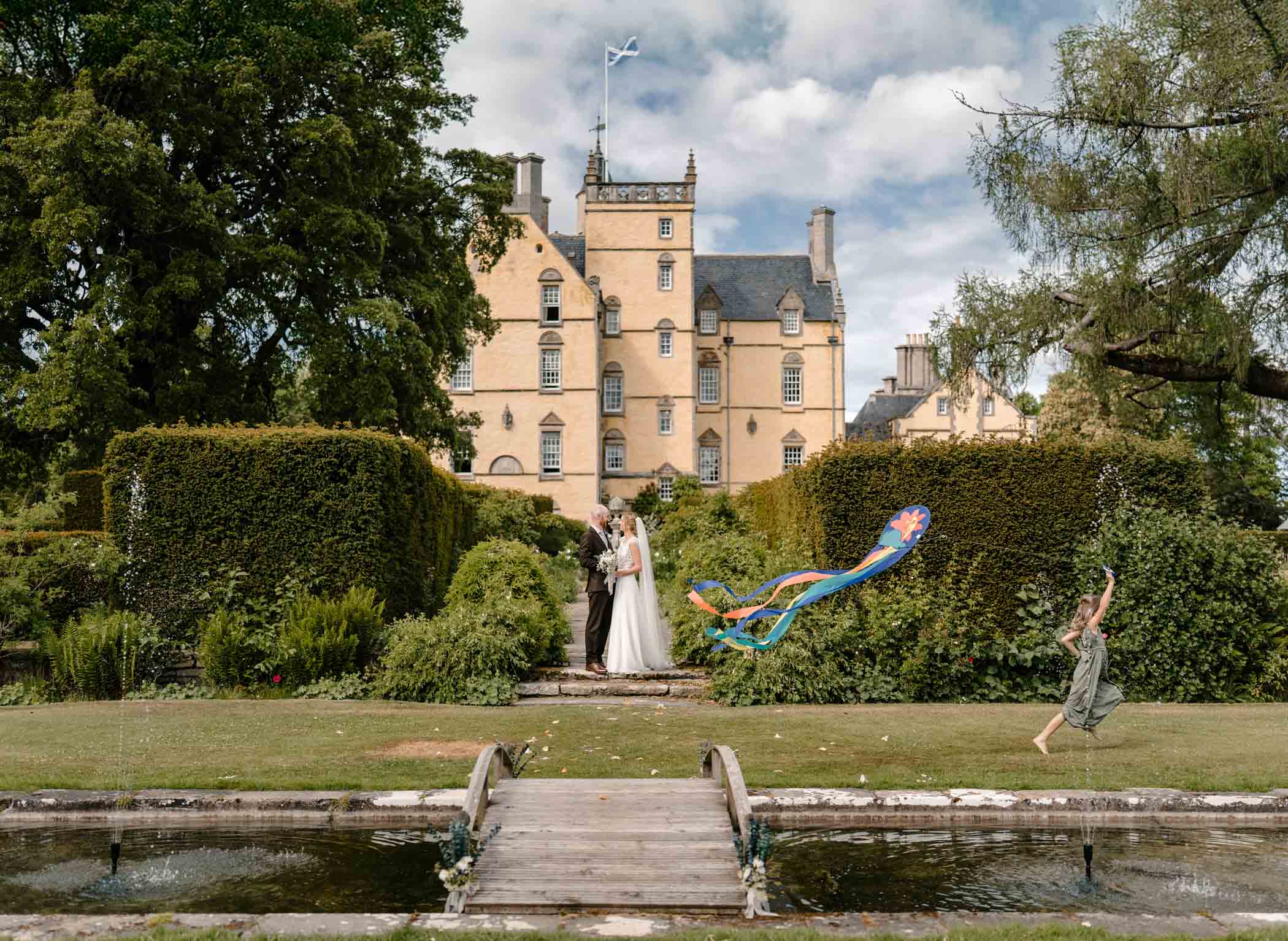 Bride and groom posing in front of Innes House as flower girl runs by flying a kite