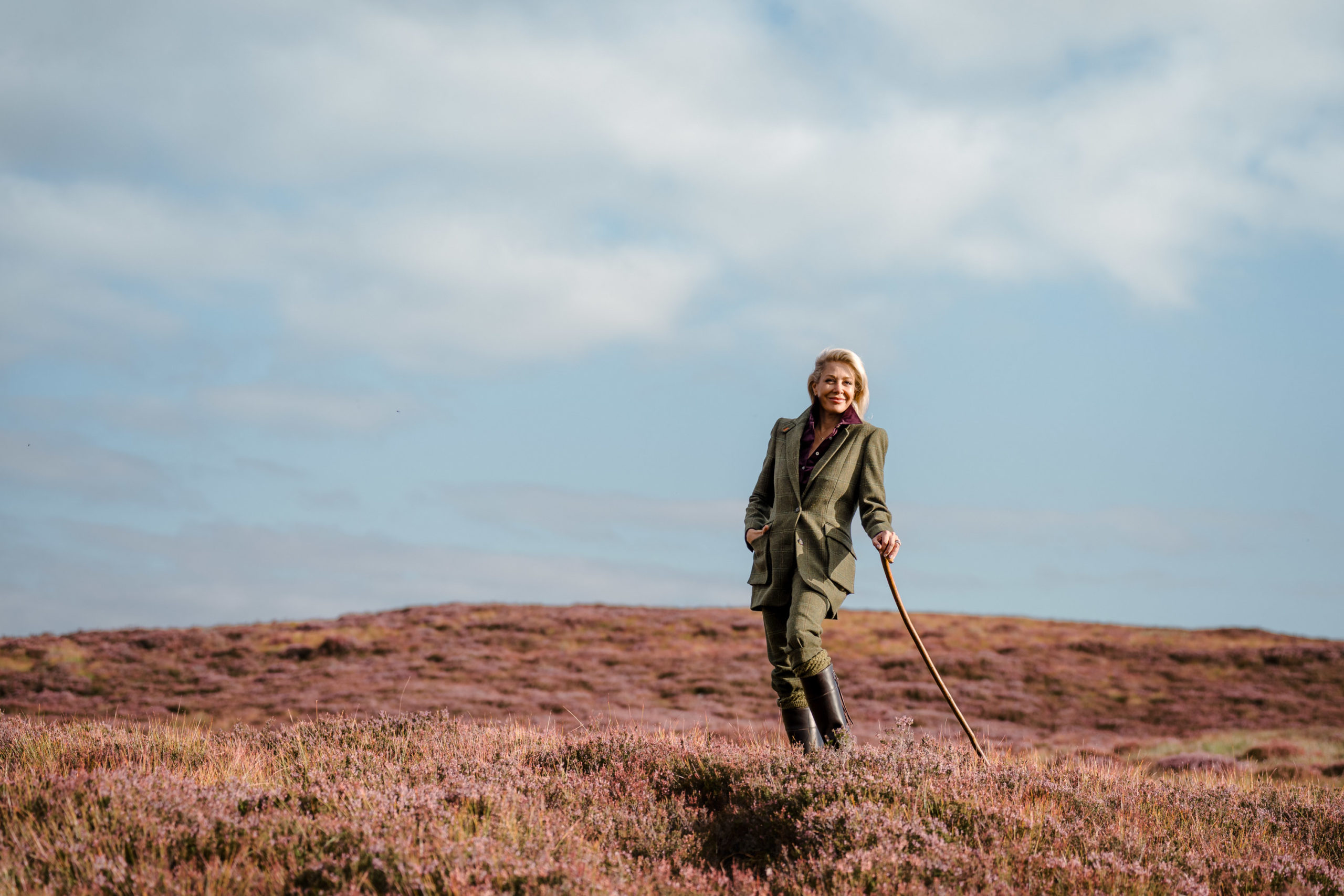 Model stands in a rural Highland landscape surrounded by purple heather, wearing a tweed jacket and matching trousers while holding a walking stick.