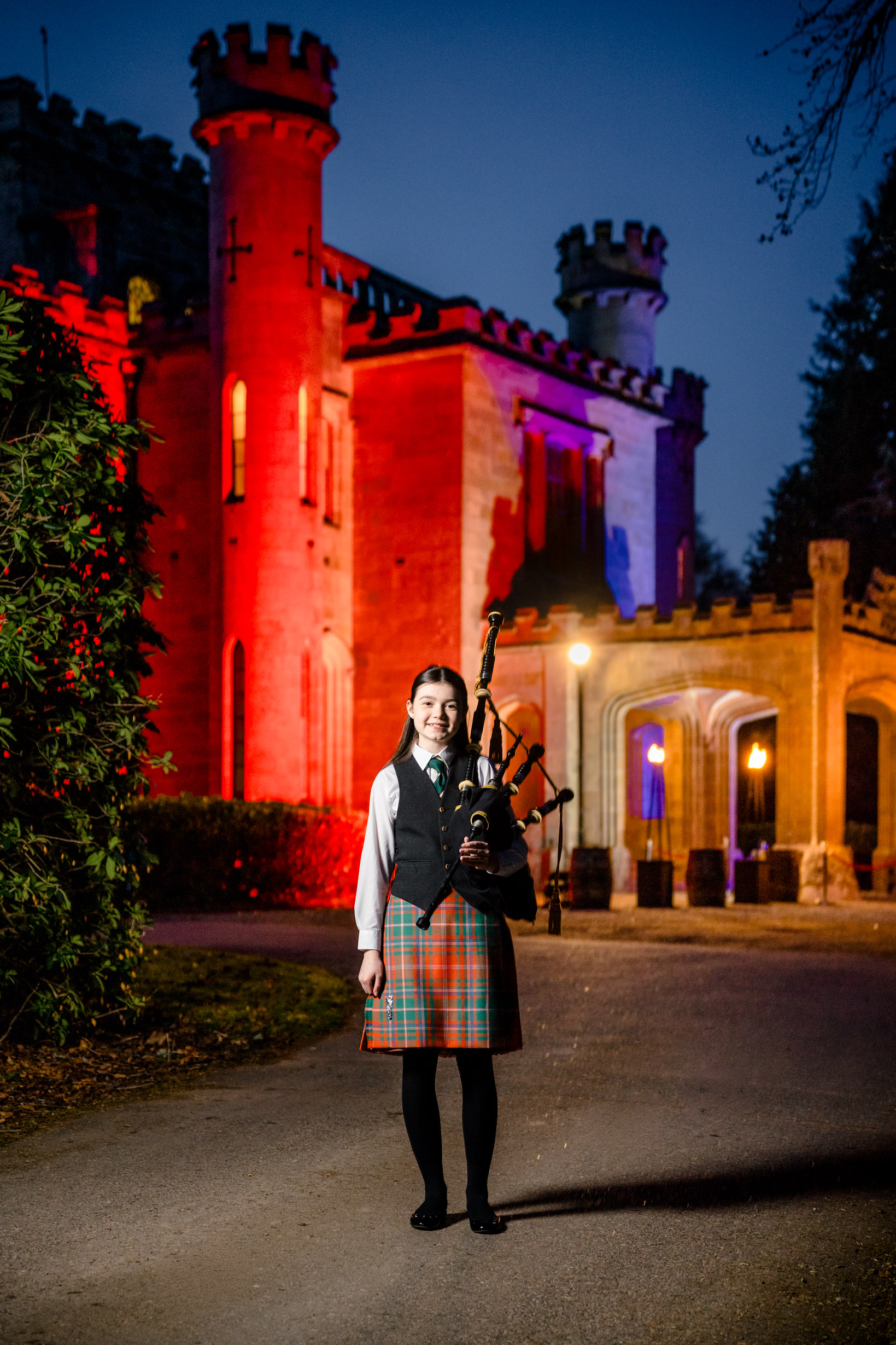 Young piper standing in front of a castle illuminated with red and blue lights, creating a striking evening portrait.