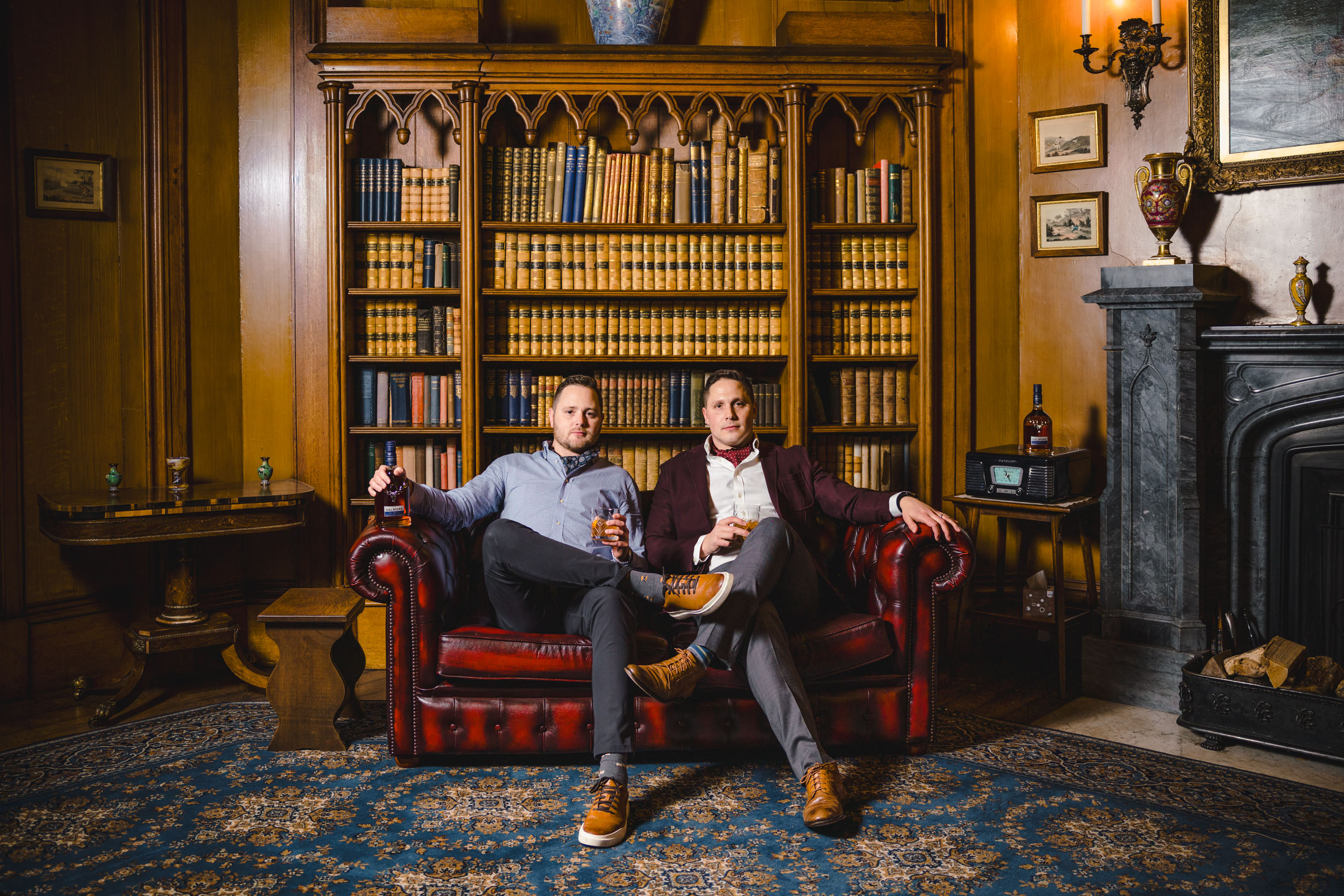 Two men sitting on a Chesterfield sofa, enjoying glasses of whisky and posing confidently in a stylish interior.