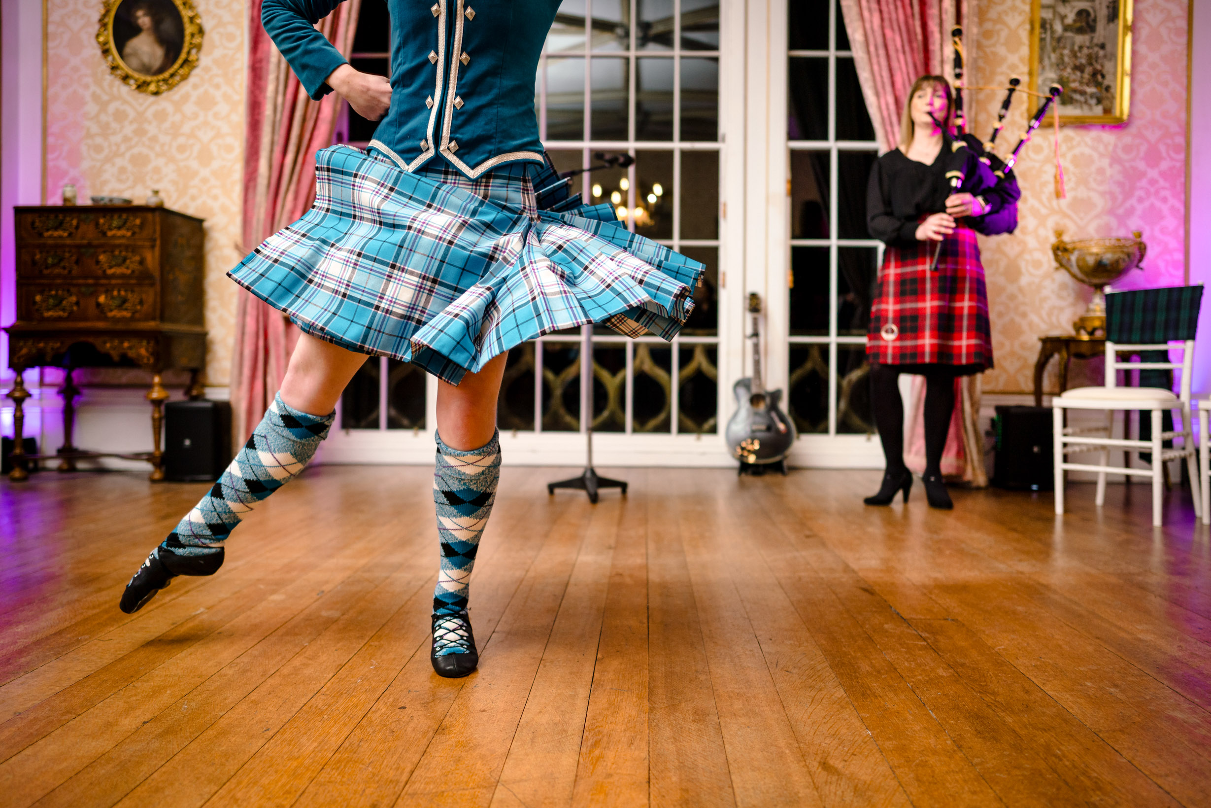 Highland dancer captured mid-performance with a kilt swirling through the air, while a piper performs in the background.