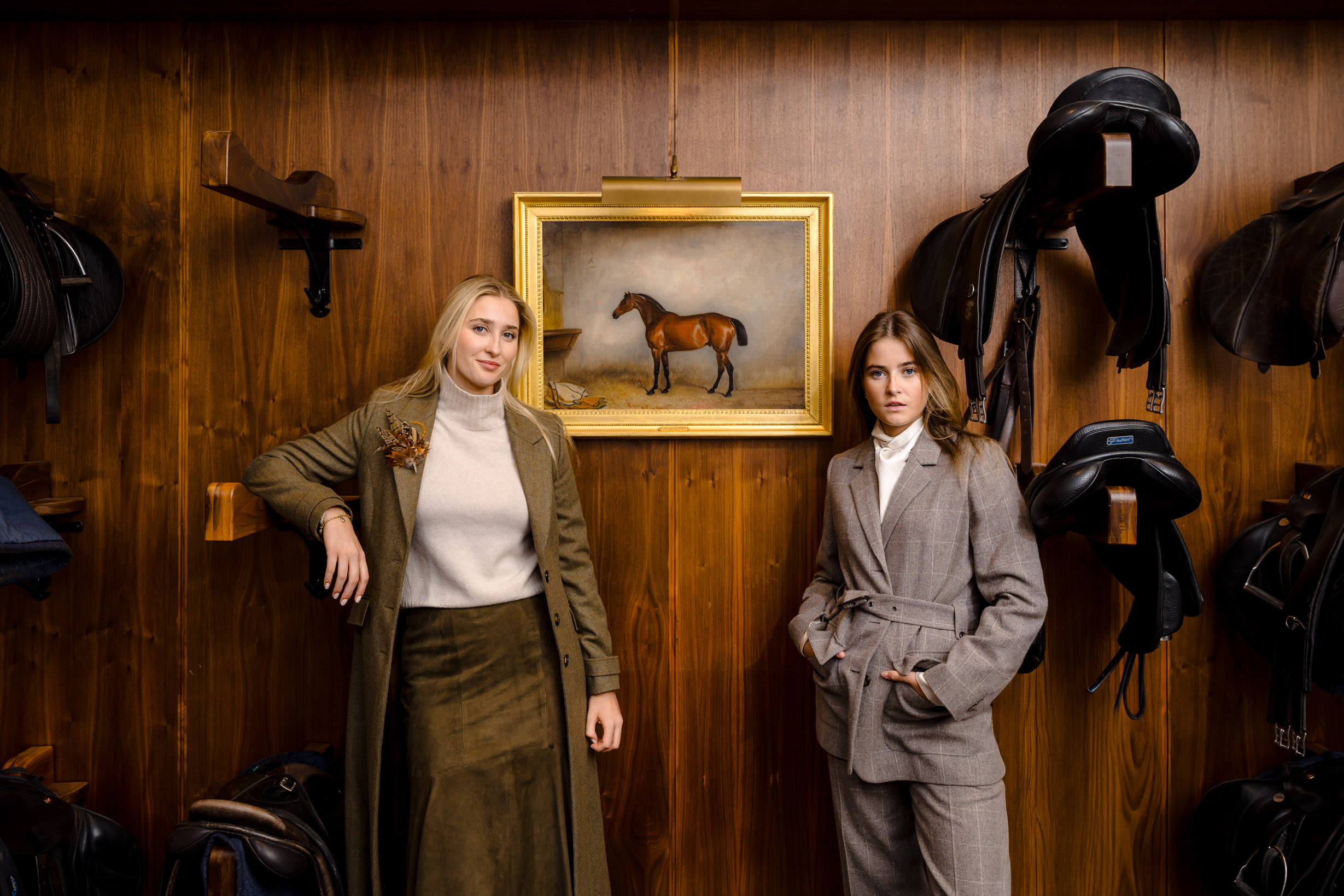 Two models posing together in elegant stables beside a painting of a horse, wearing country-style clothing