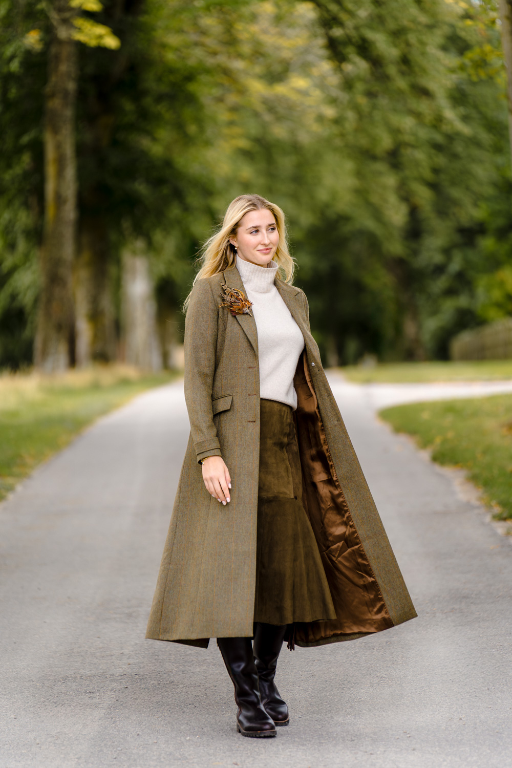 Model posing on a tree-lined street, wearing a long coat and standing among the natural surroundings in soft outdoor light.